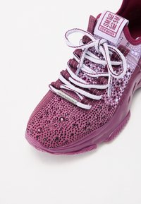 Chaussure de sport violette avec une tige texturée présentant des motifs découpés et des ornements. Lacets blancs et logo sur la languette.
