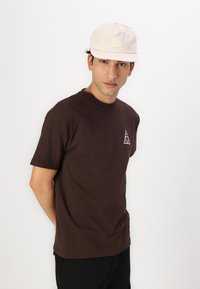 Camiseta de algodón marrón con un pequeño logo triangular blanco en el lado izquierdo del pecho, combinada con una gorra beige que tiene un parche de logo cuadrado.