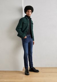 Chaqueta texturizada verde con dos bolsillos frontales, suéter de cuello alto negro, jeans azules ajustados y zapatos negros de cordones, de pie sobre un suelo de madera.