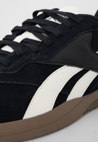 Fekete velúr sportcipő fehér bőr csíkkal, fekete kiegészítőkkel és barna gumitalppal. Jellemzője a texturált anyag és a lapos fekete fűzők.