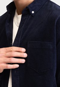 Hand die een donkerblauwe corduroy knoopshirt over een wit t-shirt met ronde hals aanpast, en de details van de borstzak en de kraag toont.