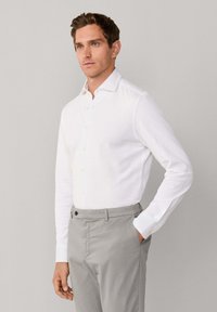 Mężczyzna ubrany w białą koszulę z długim rękawem zapinaną na guziki oraz jasnoszare spodnie, stojący z jedną ręką w kieszeni na jednolitym tle.