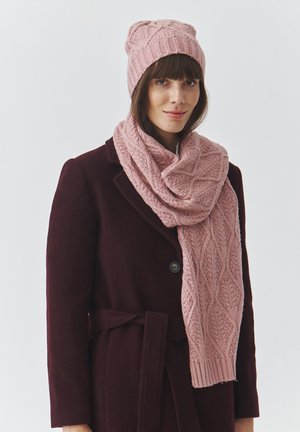 Pink knitted beanie and scarf set featuring a textured leaf pattern, paired with a dark maroon coat. Soft, warm materials.