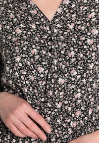 Zwarte jurk van stoffen met bloemenpatroon, met roze en witte bloemen, een v-halslijn, knopen aan de voorkant en geplooide details bij de taille.