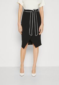 Svart omslagskjol med vit piping, hög midja och knytning. Knälång med en lätt asymmetrisk nederkant. Slät textur, kroppsnära design.