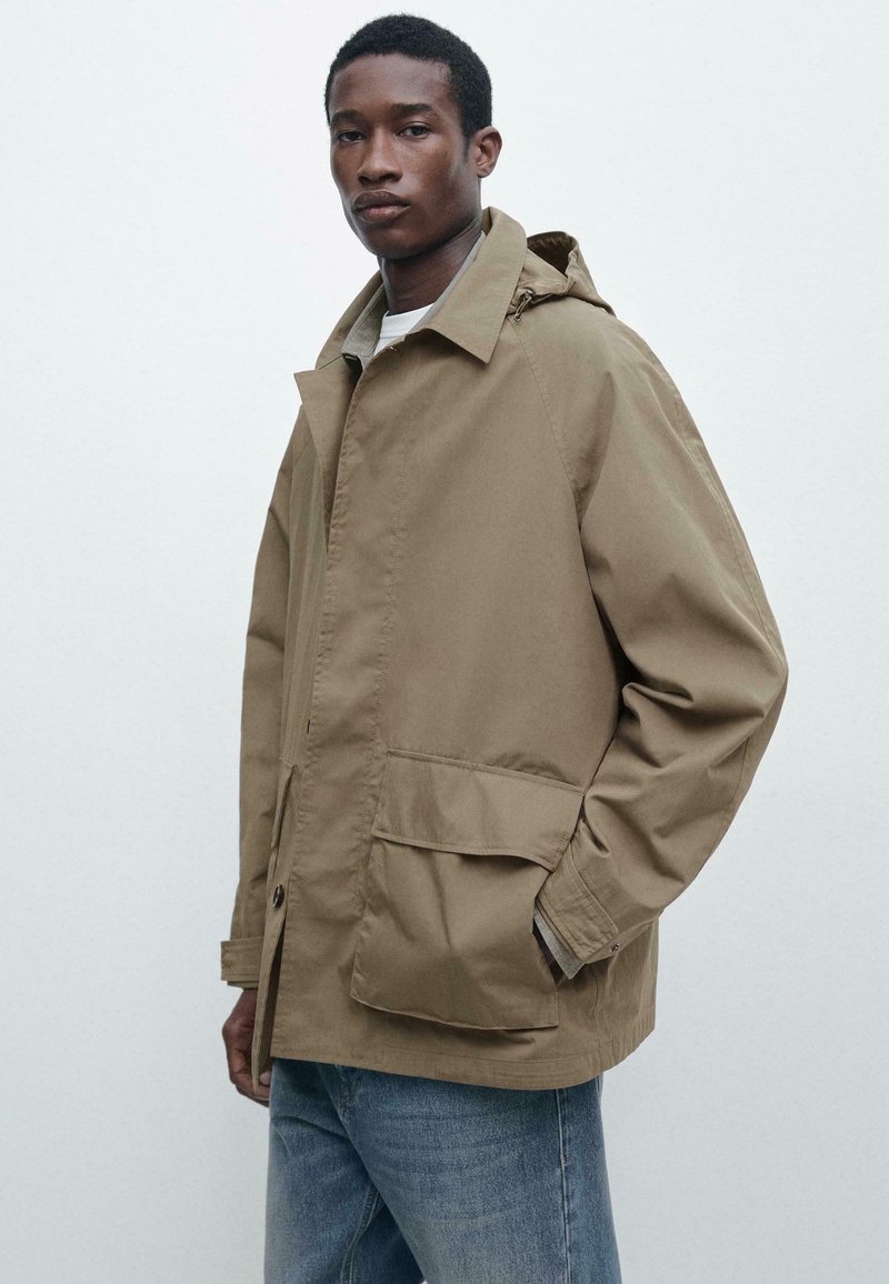 Jonge man die een lichtbruine gewatteerde jas met grote voorzakken en een capuchon draagt, gecombineerd met een blauwe spijkerbroek, staand tegen een eenvoudige lichte achtergrond.