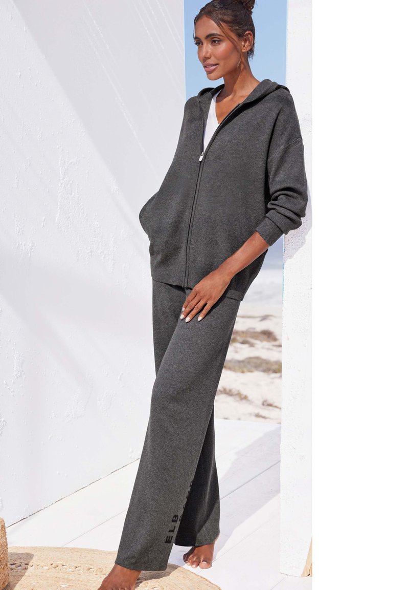 Graue Zip-Hoodie und passende Hose aus weichem Material, mit einem lässigen Schnitt, großen Taschen und dezentem Logo-Detail an der Hose.