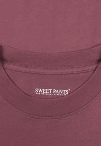 T-shirt bordeaux avec un col rond et des bords côtelés. Matière douce avec un logo visible indiquant "SWEET PANTS" mis en avant sur le col.