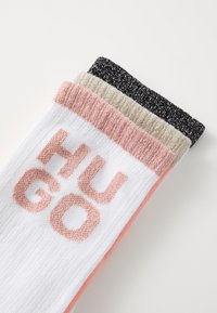 Trois paires de chaussettes : blanches avec des accents roses et le texte "HUGO", noires et beiges avec un fil métallique, dotées d'un bord côtelé.