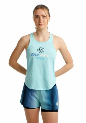 Joven mujer de pie con las manos detrás de la espalda, vistiendo una camiseta sin mangas azul claro con "Bidi Badu Padel" y pantalones deportivos de dos tonos de azul.