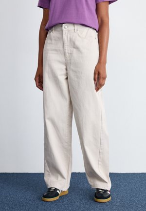 Jean large beige clair en coton avec fermeture à bouton et poches avant, associé à un t-shirt violet et des baskets noires.