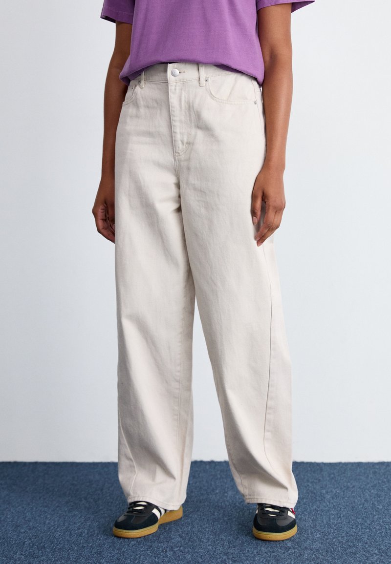 Jean large beige clair en coton avec fermeture à bouton et poches avant, associé à un t-shirt violet et des baskets noires.