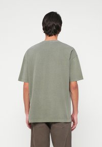 Man med mörkt hår som bär en överdimensionerad olivgrön T-shirt och bruna byxor, sedd bakifrån mot en enkel vit bakgrund.