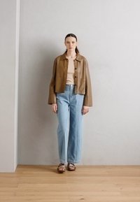 Giacca in pelle marrone corta, top beige, jeans a gamba larga blu chiaro e sandali marroni, in piedi contro un muro neutro su un pavimento di legno.