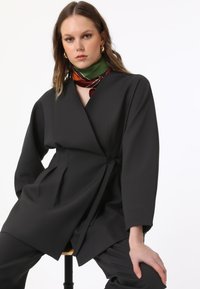 Top nero a stile avvolgente con maniche lunghe, caratterizzato da una vita con laccio e una texture morbida. Accessoriato con una sciarpa di seta multicolore attorno al collo.