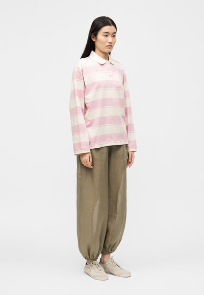 Γυναίκα που φοράει ροζ και λευκό ριγέ μακρυμάνικο πόλο, φαρδιά ελιά πράσινα παντελόνια με ελαστικές μπατζάκια και μπεζ αθλητικά παπούτσια, στέκεται ακίνητη.