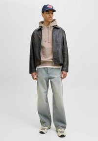 Chaqueta de cuero marrón sobre una sudadera beige, combinada con unos vaqueros de pierna ancha de color azul claro y zapatillas beige. Lleva una gorra oscura con logo.