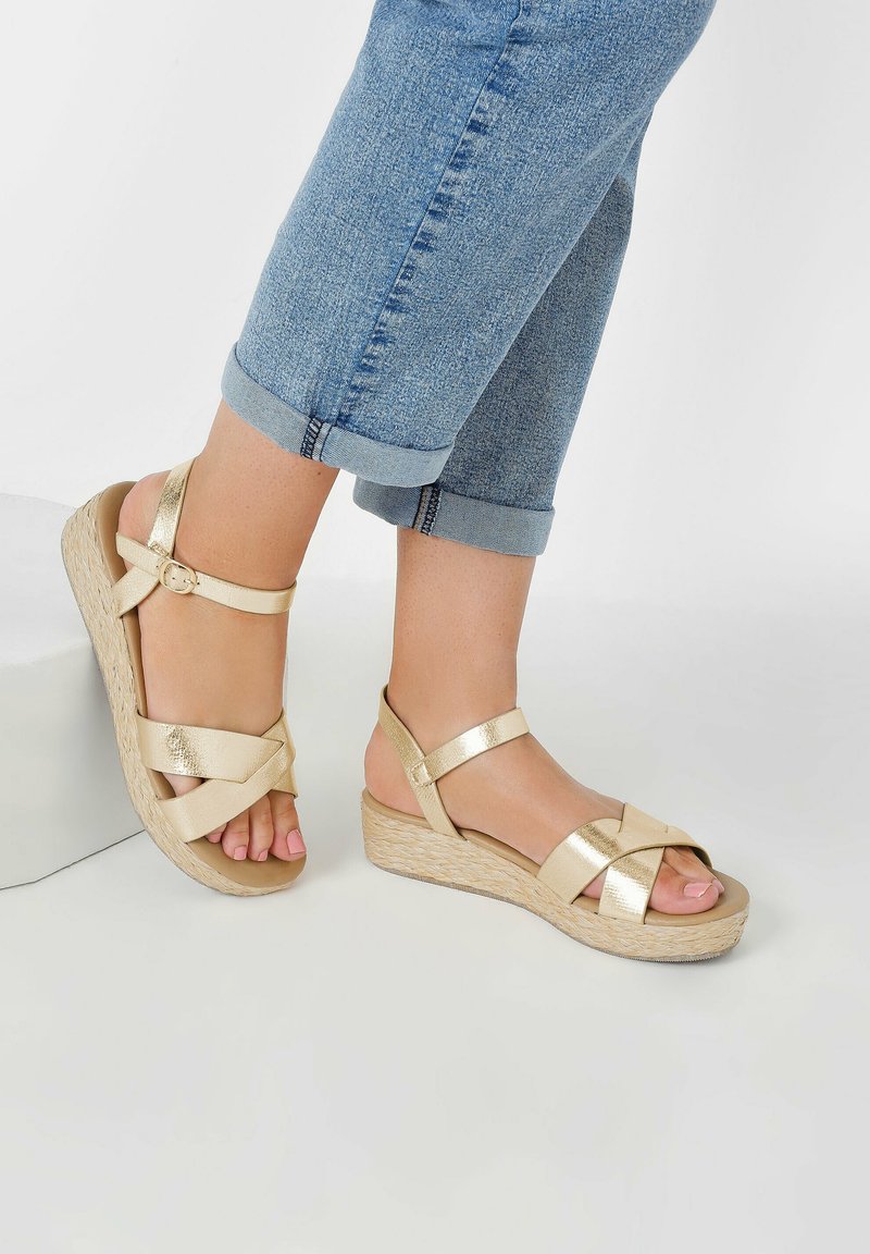 Sandales en or avec une finition métallique texturée, comportant des sangles croisées et une bride de cheville à boucle, accompagnées de jeans en denim bleu retroussés.