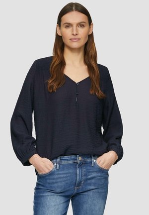 Tumme sininen, teksturoitu pitkähiha blouse V-kaula-aukolla ja nappidetaililla, yhdistettyinä sinisiin farkkuihin, kädet taskuissa.