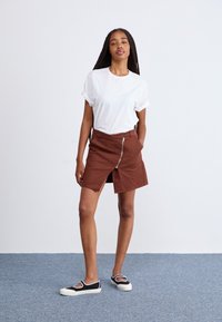Saia castanha com fecho éclair e uma fenda frontal, combinada com uma t-shirt branca de mangas dobradas e sapatos pretos com solas brancas e recortes.