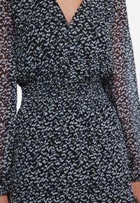 Mustat kuviolliset mekko, jossa on V-pääntie, nappiyksityiskohtia ja tiukka vyötärö; läpinäkyvät pitkät hihat, joissa on teksturoitu, abstrakti valkoinen kuvio.