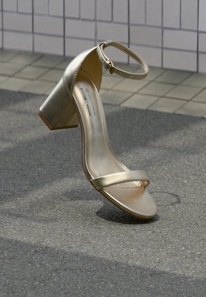 Goldene vegane Ledersandale mit Blockabsatz, Knöchelriemen und minimalistischem Riemendesign über dem Fuß. Glatte Textur und glänzendes Finish.