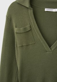 Gros plan d'un pull en maille côtelée vert olive avec un col en V, une petite patte de poche sur la poitrine gauche et un col plié.