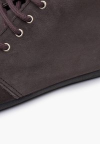 Zapato de ante marrón oscuro con punta redonda, cordones marrones y ojales plateados. Presenta una parte superior texturizada y una suela plana negra.
