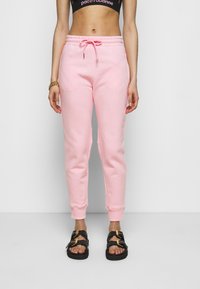 Rosa bomullssweatpants med dragsko i midjan, åtsittande ärmar och fickor, kombinerat med svarta sandaler med guldfärgade detaljer.