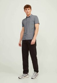 Jovem em pé, vestido com uma camisola polo cinza de mangas curtas, calças pretas e tênis de corrida cinzas, contra um fundo claro e simples.