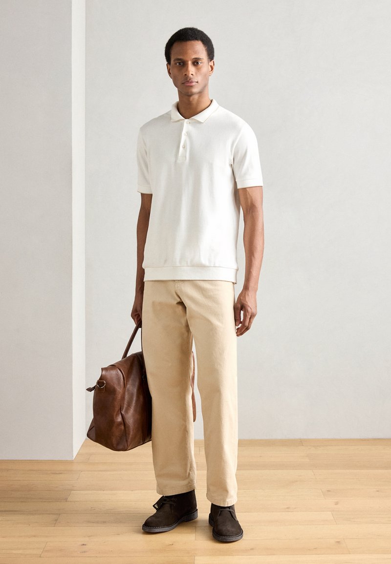 Witte polo, beige broek, bruin leren tas en donkerbruine laarzen. Eenvoudig ontwerp, gladde stof, relaxte pasvorm.
