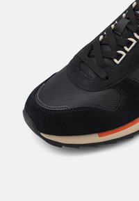 Schwarzer Sneaker mit strukturiertem Obermaterial, Wildleder-Applikationen, schwarzen Schnürsenkeln und einer beigen Zwischensohle, die durch einen orangefarbenen Streifen akzentuiert wird.