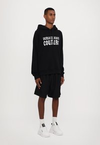 Μαύρη κουκούλα με λευκό κείμενο "Versace Jeans Couture", συνδυασμένη με μαύρες βερμούδες και λευκά αθλητικά παπούτσια. Απαλή ύφανση και χαλαρή εφαρμογή.