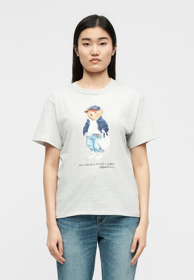 Νεαρή γυναίκα που φοράει ανοιχτό γκρι μπλουζάκι με εικονογραφημένο αρκουδάκι ντυμένο με καθημερινά ρούχα και το κείμενο "Polo Bear by Ralph Lauren," συνδυασμένο με μπλε τζιν παντελόνι.