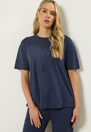 Blondynka o falowanych włosach, ubrana w luźną granatową koszulę z krótkim rękawem i pasujące spodnie, stojąca na tle białej ściany.