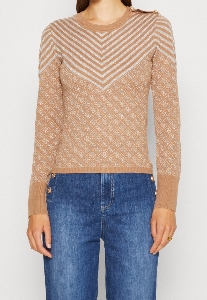 Femme portant un pull en maille beige à motifs avec des rayures en chevrons en haut, des boutons dorés aux épaules et aux poignets, associé à un jean bleu.