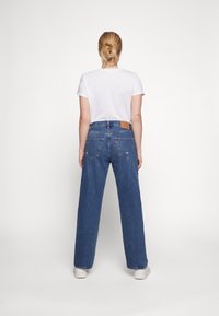 Vaqueros azules de cintura alta y pierna ancha con una textura de denim claro y una etiqueta de cuero marrón en la cinturilla trasera. Combinados con una camiseta blanca básica.