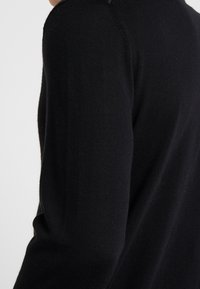Maglione nero lavorato a maglia che mostra un primo piano della spalla e della cucitura della manica. Il tessuto appare liscio con una leggera texture. Nessun motivo o accento visibile.