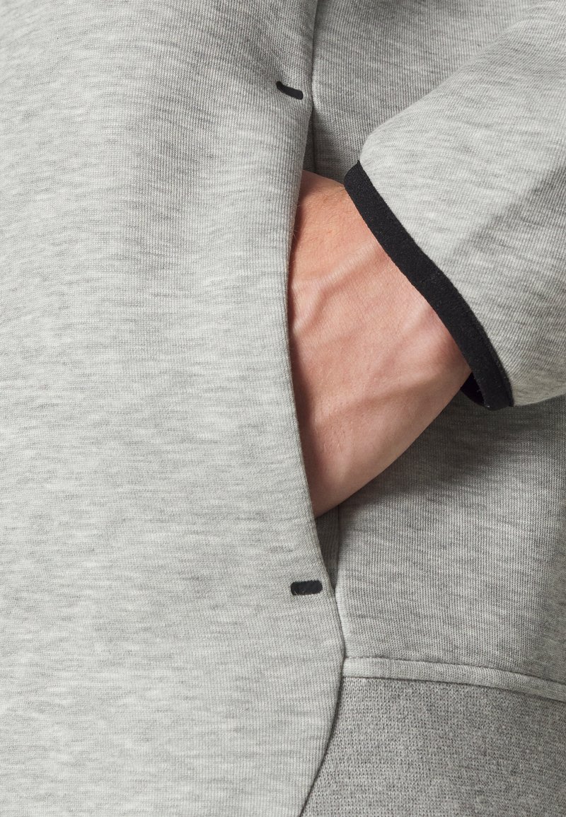 Sweatshirt gris avec une finition texturée. Offre une poche avant et des accents noirs au niveau des poignets. Le tissu semble doux et décontracté.