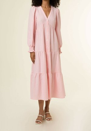 Kvinde iført en lang, lyserød kjole med knapper, puffede ærmer og rynkede manchetter, kombineret med hvide åbne sandaler med hæl.