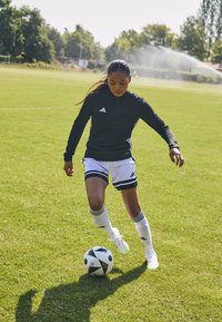 Svart langermet treningstopp med hvite detaljer, hvite shorts med svarte striper, og en svart-hvitt fotball på grønt gress.