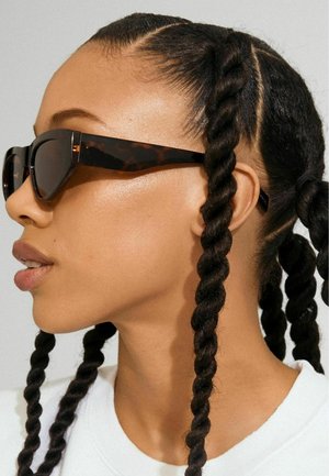 Occhiali da sole tartarugati con un design geometrico, caratterizzati da lenti scure e montature robuste. Capelli acconciati in trecce intrecciate lungo il viso.