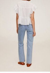 Biały top z krótkim rękawem z falbankami na krawędziach, teksturowany tył, w połączeniu z jasnoniebieskimi jeansami o prostych nogawkach i brązowymi sandałami. Neutralne tło.