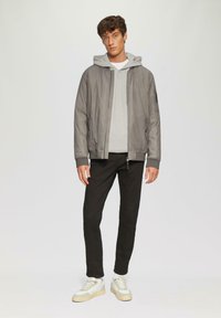 Chaqueta bomber ligera y gris con puños de canalé, sudadera gris debajo, pantalones negros y zapatillas beige, con cremalleras y bolsillos.