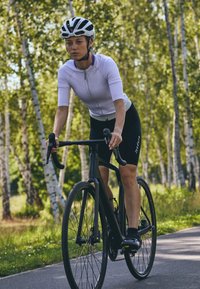 Cyclist on a black road bike, wearing a white short-sleeve jersey and black shorts, with a black and white helmet, on a wooded path.