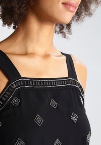 Haut noir sans manches avec un col orné de perles et des motifs géométriques en sequins, présentant une texture de tissu lisse et une coupe ajustée.