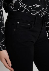 Jeans de denim negro con una forma ajustada, que presentan una cinturilla visible, cierres de botón y un detalle de logotipo. Complementados con una blusa de patrones en blanco y negro.