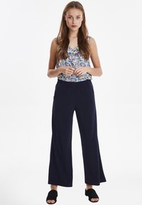 Ärmelloses Top mit Blumenmuster in Blau und Gelb; marineblaue weite Hose; Modell trägt schwarze Slippers; Hände vorne zusammengelegt.