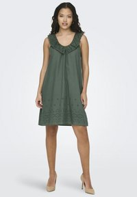 Zelena haljina bez rukava s volanima oko vrata, vezenim okomitim uzorkom na rubu i labavim krojem. Tkanina izgleda glatko i lagano.