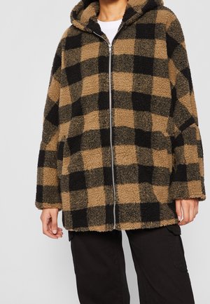 Bruine en zwarte geruite fleece-hoodie met rits, gedragen over een wit overhemd met een zwarte broek.